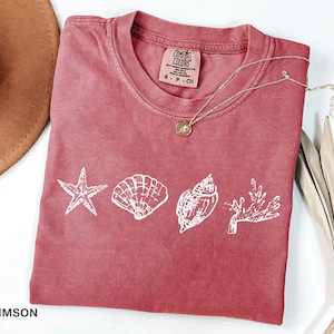 Op de afbeelding: Een koraalrood T-shirt met een witte afbeelding van een zeester, schelpen en koraal. Het shirt heeft een label met de tekst "COMFORT COLORS" en "S - P - CH".