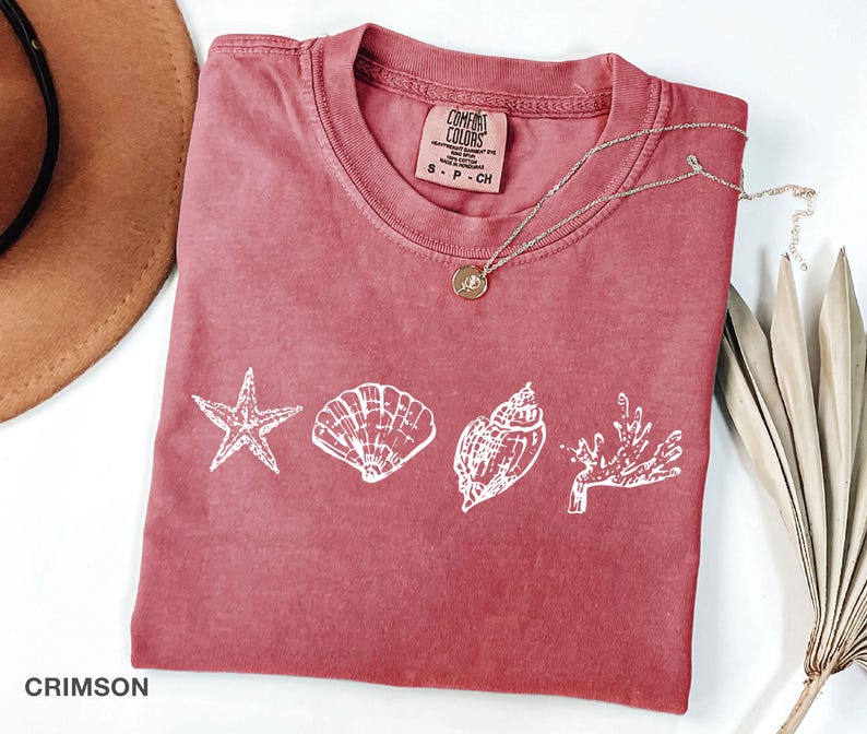 Op de afbeelding: Een koraalrood T-shirt met een witte afbeelding van een zeester, schelpen en koraal. Het shirt heeft een label met de tekst "COMFORT COLORS" en "S - P - CH".