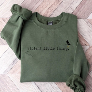 以下が含まれることがあります： 「violent little thing.」というテキストと小さな黒いドラゴンのシルエットが描かれた緑色のスウェットシャツです。