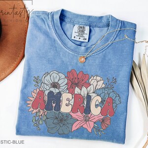 May include: A blue t-shirt with a vintage-style graphic of the word "AMERICA" in colorful letters surrounded by flowers. The shirt is made of a soft, comfortable material and has a distressed look.