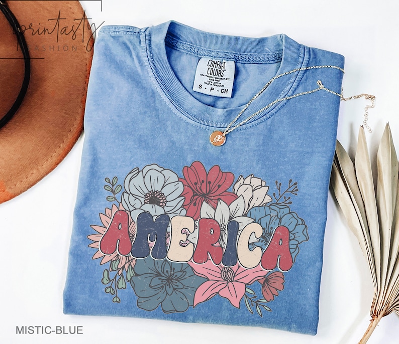 May include: A blue t-shirt with a vintage-style graphic of the word "AMERICA" in colorful letters surrounded by flowers. The shirt is made of a soft, comfortable material and has a distressed look.