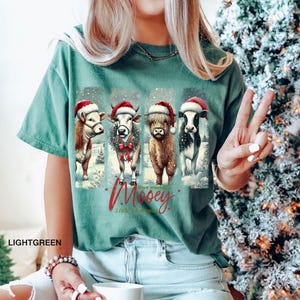 May include: Light green t-shirt featuring four cows wearing Santa hats and standing in a snowy scene. The text "Have yourself a Mooey Little Christmas" is printed in red script. The shirt is a comfortable, casual style.
