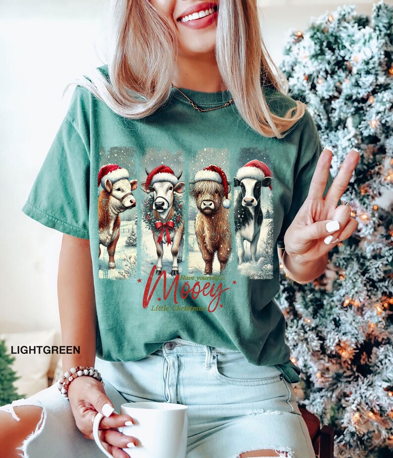 May include: Light green t-shirt featuring four cows wearing Santa hats and standing in a snowy scene. The text "Have yourself a Mooey Little Christmas" is printed in red script. The shirt is a comfortable, casual style.