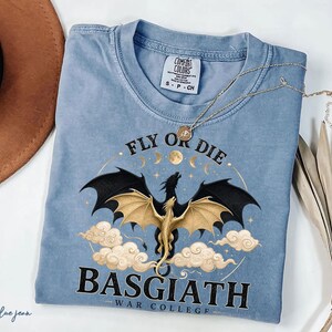 May include: A blue jean coloured t-shirt featuring a dragon graphic. The dragon has black wings and a gold body, surrounded by clouds and the text "FLY OR DIE BASGIATH WAR COLLEGE". A gold necklace rests on the shirt.