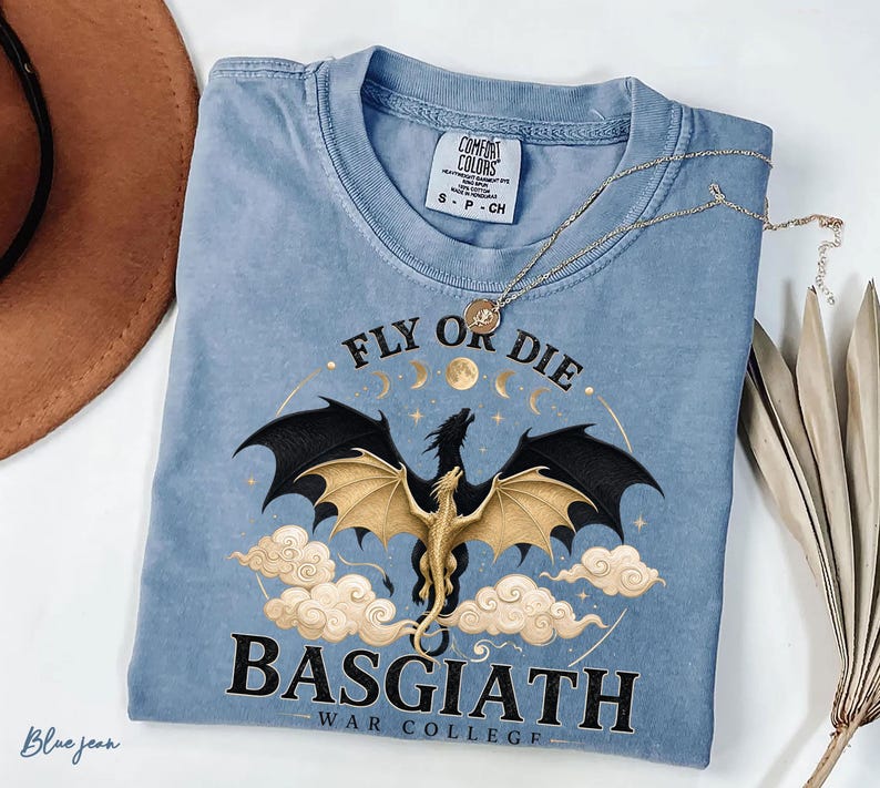 May include: A blue jean coloured t-shirt featuring a dragon graphic. The dragon has black wings and a gold body, surrounded by clouds and the text "FLY OR DIE BASGIATH WAR COLLEGE". A gold necklace rests on the shirt.