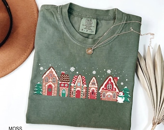 May include: Moss green Comfort Colors t-shirt featuring a festive gingerbread house design with red, white, and brown details. The shirt includes a necklace and a tag that reads "Comfort Colors". The word "MOSS" is printed at the bottom.