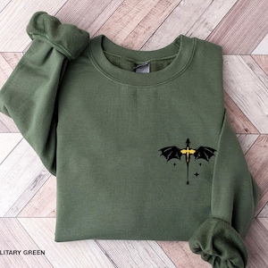 Puede incluir: Una sudadera de color verde militar con un gr&aacute;fico de drag&oacute;n negro con detalles amarillos. El drag&oacute;n est&aacute; volando con las alas extendidas. El drag&oacute;n est&aacute; rodeado de peque&ntilde;as estrellas.