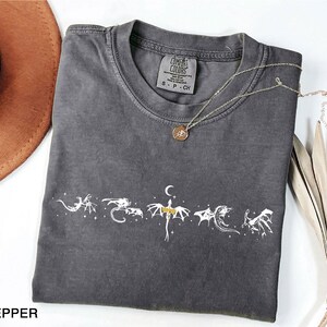 May include: A dark gray t-shirt with a white dragon graphic design. The shirt has a crew neck and short sleeves. A gold necklace with a pendant is draped across the shirt. The word "PEPPER" is at the bottom left.