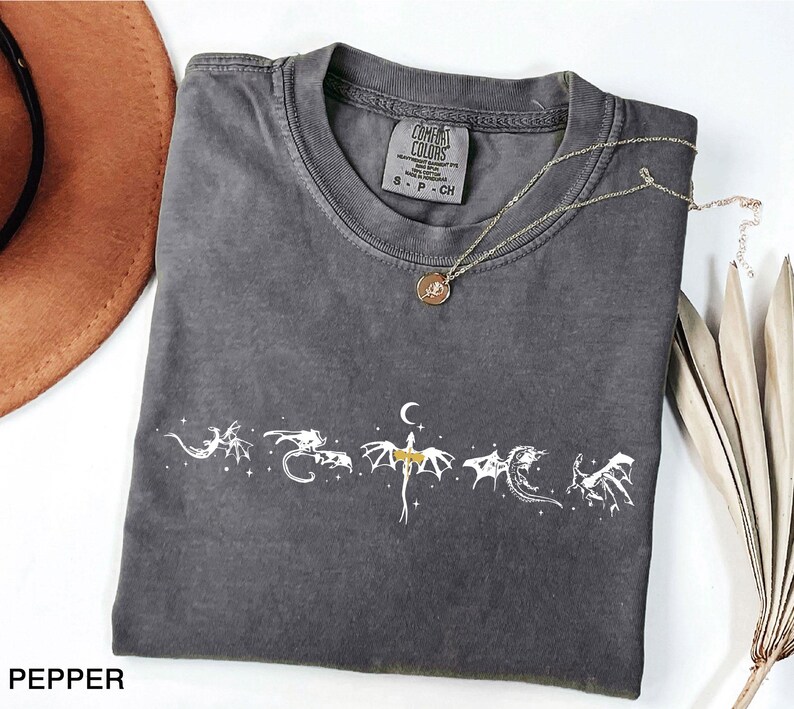 May include: A dark gray t-shirt with a white dragon graphic design. The shirt has a crew neck and short sleeves. A gold necklace with a pendant is draped across the shirt. The word "PEPPER" is at the bottom left.