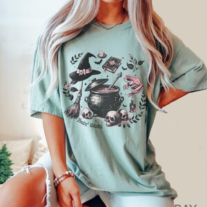 May include: A light green t-shirt with a graphic print of a witch's cauldron, a broom, a frog, a hat, and skulls. The text "Feelin' Witchy" is printed below the graphic.