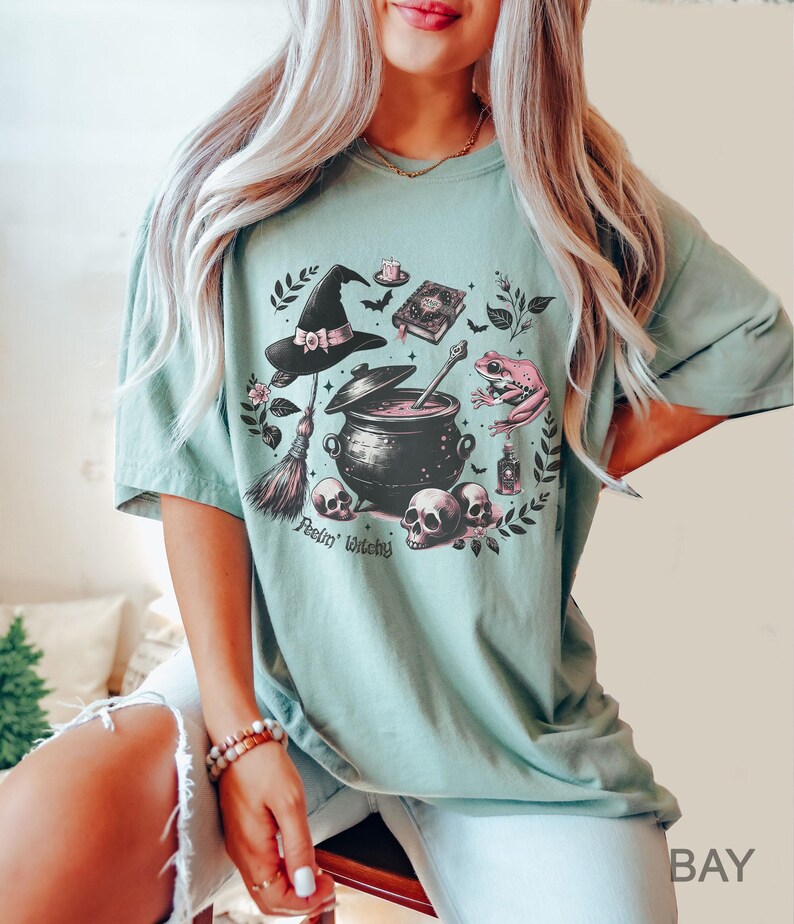 May include: A light green t-shirt with a graphic print of a witch's cauldron, a broom, a frog, a hat, and skulls. The text "Feelin' Witchy" is printed below the graphic.