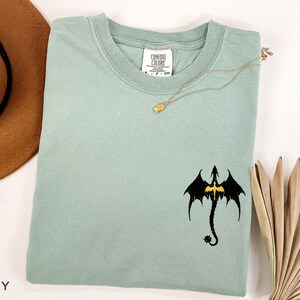 May include: A light blue t-shirt with a black dragon graphic. The dragon has a yellow outline and is facing forward with its wings spread. The shirt has a crew neck and short sleeves.