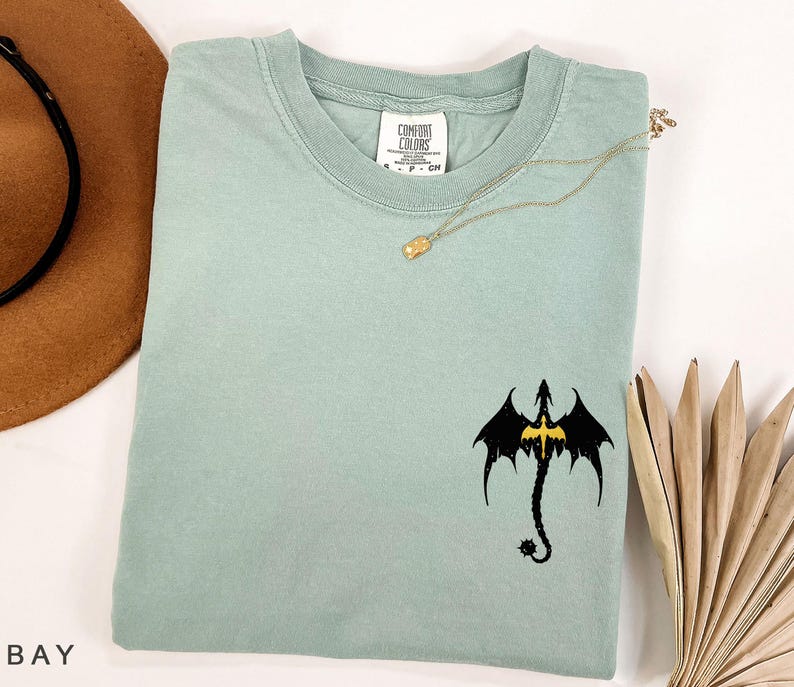 May include: A light blue t-shirt with a black dragon graphic. The dragon has a yellow outline and is facing forward with its wings spread. The shirt has a crew neck and short sleeves.