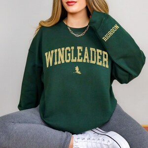 May include: A dark green sweatshirt with the text "WINGLEADER" in gold lettering and a small dragon graphic. The sleeve has the text "RIDORSON" in gold lettering.