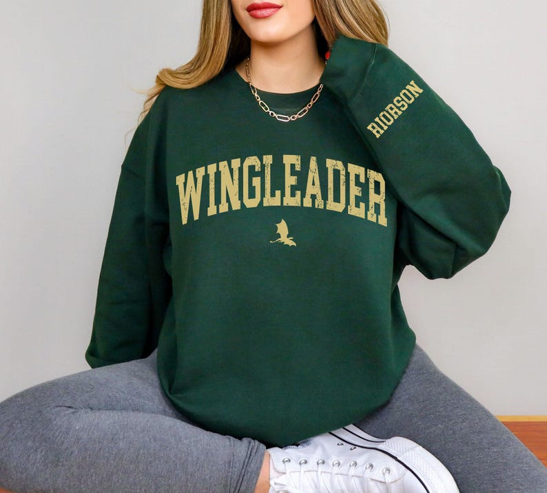 May include: A dark green sweatshirt with the text "WINGLEADER" in gold lettering and a small dragon graphic. The sleeve has the text "RIDORSON" in gold lettering.