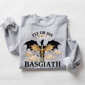 May include: A light gray sweatshirt featuring a dragon design with the text "FLY OR DIE" above and "BASGIATH WAR COLLEGE" below. The dragon has black and gold wings, with a full moon and clouds in the background.