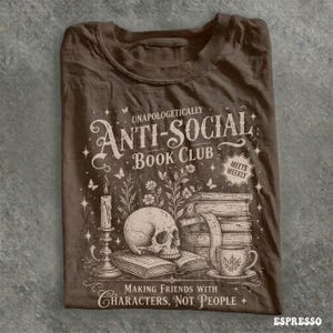 May include: Espresso-colored t-shirt featuring the text "Unapologetically Anti-Social Book Club" with a skull, books, candle, and teacup graphic. The shirt also includes the phrase "Making Friends With Characters, Not People."