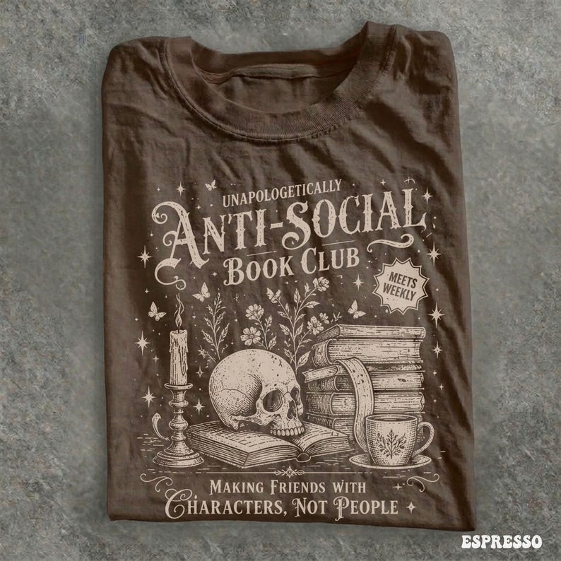 May include: Espresso-colored t-shirt featuring the text "Unapologetically Anti-Social Book Club" with a skull, books, candle, and teacup graphic. The shirt also includes the phrase "Making Friends With Characters, Not People."