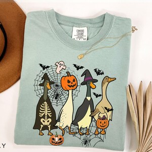 Puede incluir: Camiseta Comfort Colors verde claro con un dise&ntilde;o de Halloween. El dise&ntilde;o incluye cuatro patos disfrazados, una calabaza, un fantasma, murci&eacute;lagos y una telara&ntilde;a. La camiseta tiene cuello redondo y mangas cortas.