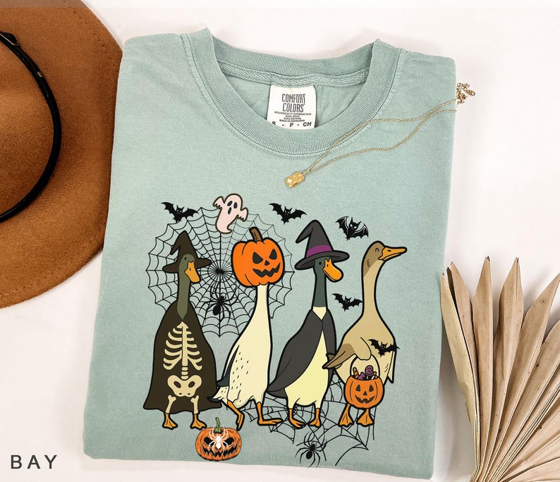 Puede incluir: Camiseta Comfort Colors verde claro con un dise&ntilde;o de Halloween. El dise&ntilde;o incluye cuatro patos disfrazados, una calabaza, un fantasma, murci&eacute;lagos y una telara&ntilde;a. La camiseta tiene cuello redondo y mangas cortas.