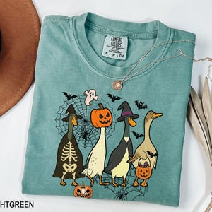 Puede incluir: Camiseta verde claro con un dise&ntilde;o de Halloween. El gr&aacute;fico muestra cuatro patos de dibujos animados disfrazados para Halloween, incluyendo un esqueleto, una bruja y una calabaza. El dise&ntilde;o tambi&eacute;n presenta una telara&ntilde;a, murci&eacute;lagos y un fantasma. Un collar plateado est&aacute; sobre la camiseta.