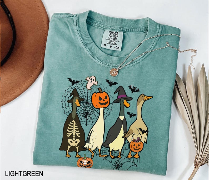 Puede incluir: Camiseta verde claro con un dise&ntilde;o de Halloween. El gr&aacute;fico muestra cuatro patos de dibujos animados disfrazados para Halloween, incluyendo un esqueleto, una bruja y una calabaza. El dise&ntilde;o tambi&eacute;n presenta una telara&ntilde;a, murci&eacute;lagos y un fantasma. Un collar plateado est&aacute; sobre la camiseta.