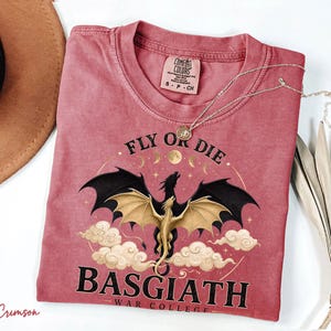 May include: Crimson t-shirt featuring a dragon design with the text "FLY OR DIE" and "BASGIATH WAR COLLEGE". The dragon has black wings and a gold body, set against clouds and moons. A silver necklace is draped across the shirt. The shirt is a Comfort Colors brand.
