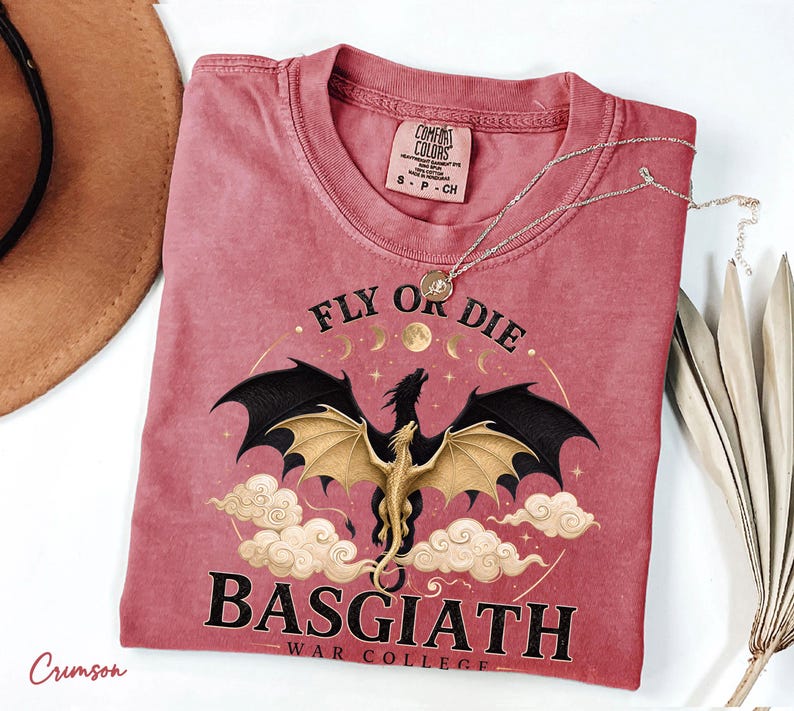 May include: Crimson t-shirt featuring a dragon design with the text "FLY OR DIE" and "BASGIATH WAR COLLEGE". The dragon has black wings and a gold body, set against clouds and moons. A silver necklace is draped across the shirt. The shirt is a Comfort Colors brand.
