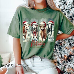 May include: Moss green t-shirt featuring a graphic of four cows wearing Santa hats and a winter scene. The text "Have yourself a Mooey Little Christmas" is below the cows. The shirt is a relaxed fit.