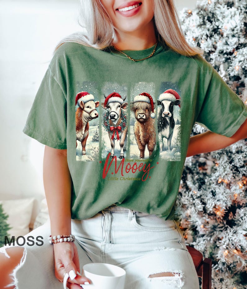 May include: Moss green t-shirt featuring a graphic of four cows wearing Santa hats and a winter scene. The text "Have yourself a Mooey Little Christmas" is below the cows. The shirt is a relaxed fit.