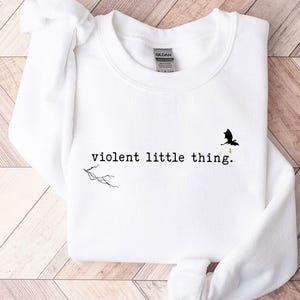 以下が含まれることがあります： 白いスウェットシャツに「violent little thing」という文字がプリントされています。テキストの上に小さな黒いドラゴンのシルエットがあります。