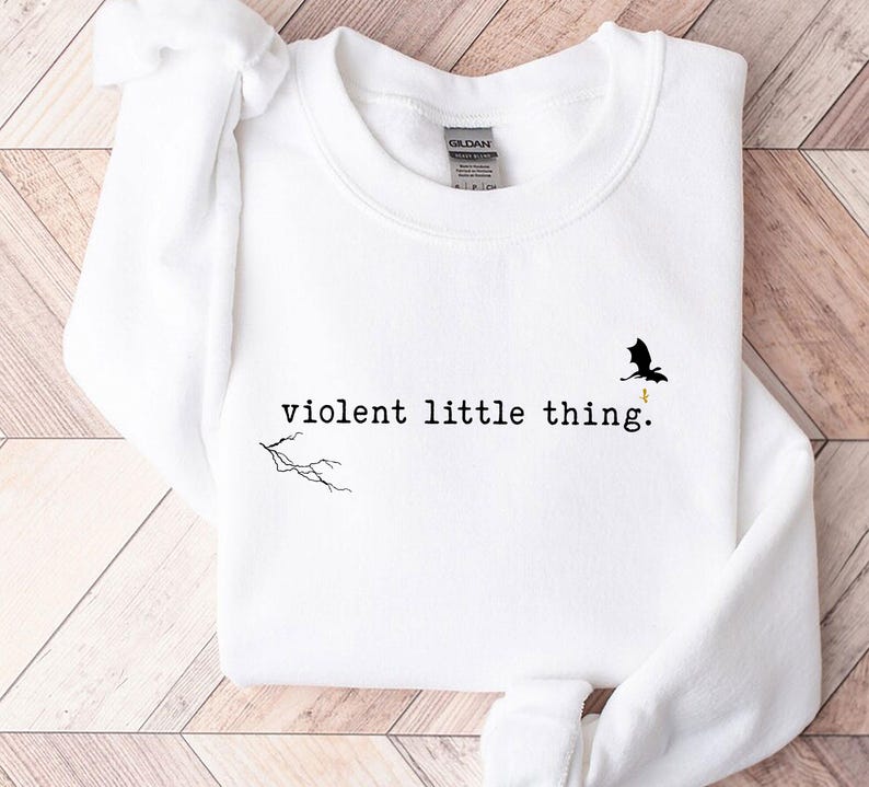 以下が含まれることがあります： 白いスウェットシャツに「violent little thing」という文字がプリントされています。テキストの上に小さな黒いドラゴンのシルエットがあります。