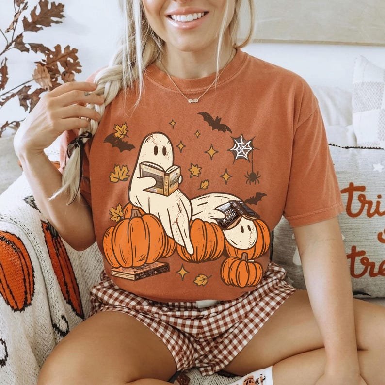 Może przedstawiać: T-shirt z kr&oacute;tkim rękawem w kolorze rdzawym z grafiką przedstawiającą dwa białe duchy czytające książki wśr&oacute;d pomarańczowych dyń. Wz&oacute;r zawiera małe nietoperze, pajęczynę i rozrzucone jesienne liście, z napisem Onyx na jednej z książek.