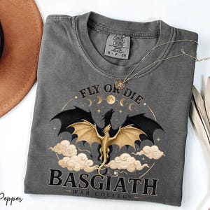 May include: A grey Comfort Colors t-shirt featuring a dragon graphic. The design includes a dragon with black wings and a gold body, set against clouds, moons, and stars. Text reads "FLY OR DIE" and "BASGIATH WAR COLLEGE."