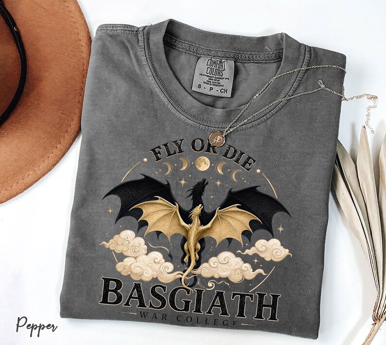 May include: A grey Comfort Colors t-shirt featuring a dragon graphic. The design includes a dragon with black wings and a gold body, set against clouds, moons, and stars. Text reads "FLY OR DIE" and "BASGIATH WAR COLLEGE."
