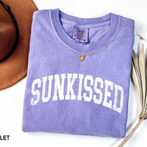 May include: A violet Comfort Colors t-shirt with the word "SUNKISSED" in white block letters. A gold necklace with a small pendant rests on the shirt. A brown hat and dried plant are in the background.