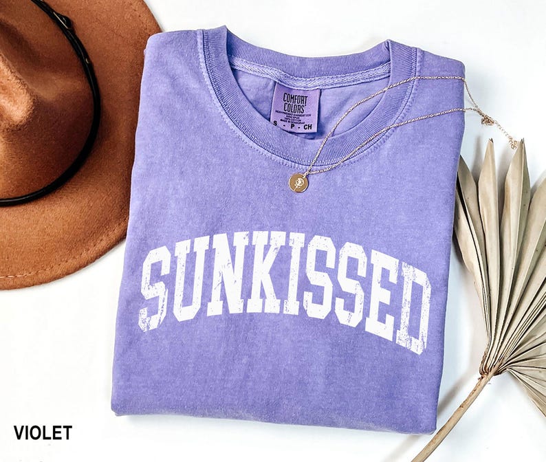 May include: A violet Comfort Colors t-shirt with the word "SUNKISSED" in white block letters. A gold necklace with a small pendant rests on the shirt. A brown hat and dried plant are in the background.