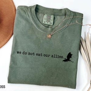 May include: A moss green t-shirt with the text "we do not eat our allies." and a black silhouette of a dragon.