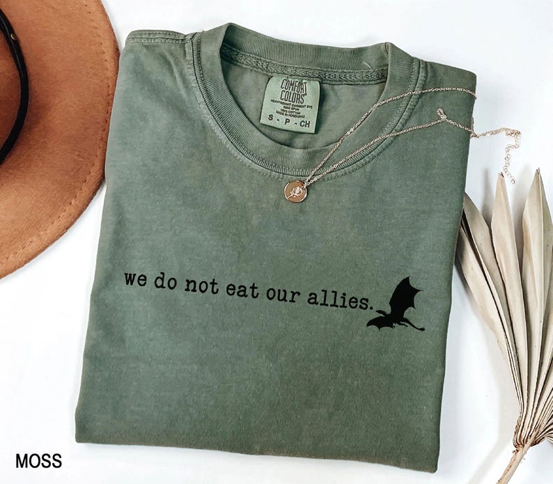 May include: A moss green t-shirt with the text "we do not eat our allies." and a black silhouette of a dragon.