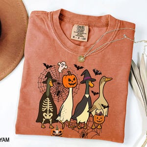 Puede incluir: Una camiseta Comfort Colors de color terracota con un gr&aacute;fico de tem&aacute;tica de Halloween. El dise&ntilde;o incluye cuatro patos de dibujos animados disfrazados para Halloween, murci&eacute;lagos, ara&ntilde;as y una telara&ntilde;a. Un collar plateado con un colgante est&aacute; sobre la camiseta.