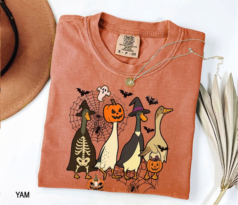 Puede incluir: Una camiseta Comfort Colors de color terracota con un gr&aacute;fico de tem&aacute;tica de Halloween. El dise&ntilde;o incluye cuatro patos de dibujos animados disfrazados para Halloween, murci&eacute;lagos, ara&ntilde;as y una telara&ntilde;a. Un collar plateado con un colgante est&aacute; sobre la camiseta.