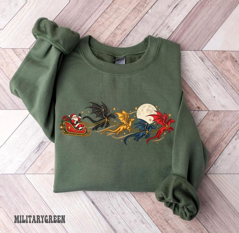 May include: An olive green crewneck sweatshirt featuring a graphic print of Santa Claus in a red sleigh pulled by four flying dragons in black, gold, blue, and red colors, set against a full moon and small yellow stars.