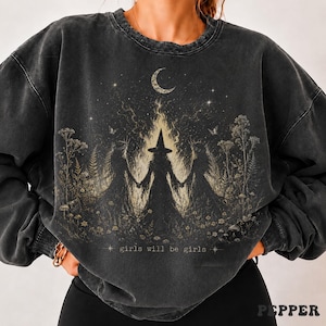 May include: A charcoal gray crewneck sweatshirt featuring a graphic print of three figures in pointed hats holding hands amidst tall wildflowers and flames. Above them is a crescent moon and stars. The text "girls will be girls" is printed below.