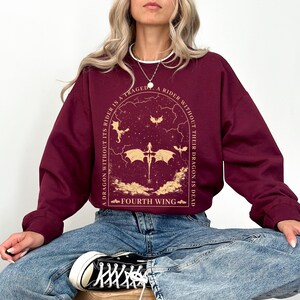 Pode incluir: Uma camisola de cor vinho com uma estampa gr&aacute;fica de um drag&atilde;o e o texto "A dragon without its rider is a tragedy. A rider without their dragon is dead. Fourth Wing."