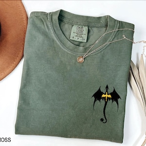 May include: A moss green t-shirt with a black dragon graphic and a yellow cross. The shirt has the text "COMFORT COLORS" and "S - P - CH" on the tag.