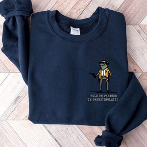 May include: Navy blue sweatshirt featuring a cartoon frog wearing a hat and holding a gun. The text "HOLD ON PARTNER I'M OVERSTIMULATED" is printed below the frog.