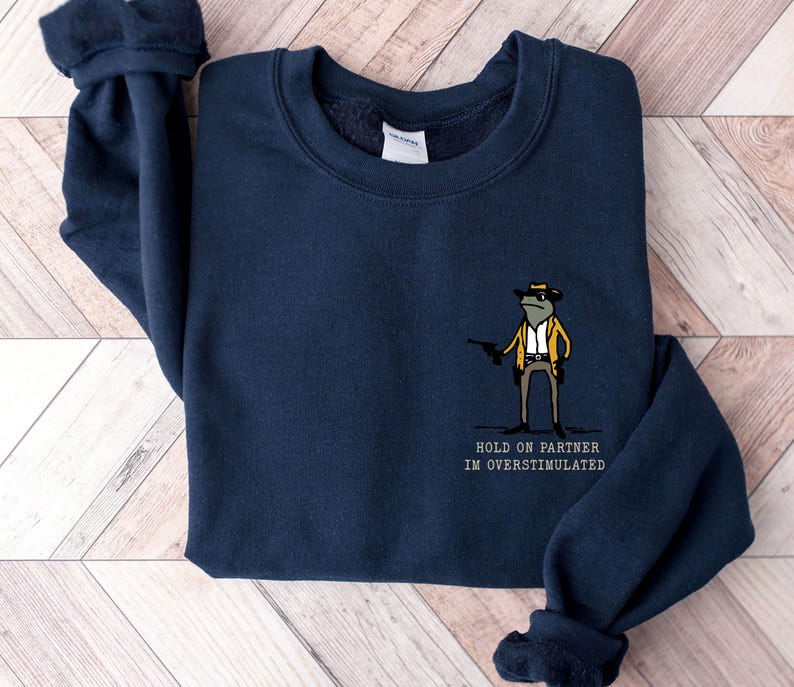 May include: Navy blue sweatshirt featuring a cartoon frog wearing a hat and holding a gun. The text "HOLD ON PARTNER I'M OVERSTIMULATED" is printed below the frog.