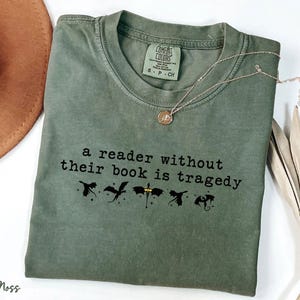 May include: A moss green Comfort Colors t-shirt with the text "a reader without their book is tragedy" in black. Below the text are black dragon silhouettes. A gold necklace with a pendant is on the shirt.