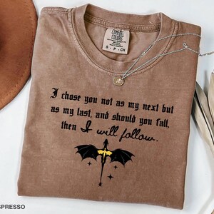 May include: Espresso-colored t-shirt with the text "I chose you not as my next but as my last, and should you fall, then I will follow." A black dragon graphic with yellow accents is below the text. A silver necklace is draped across the shirt.