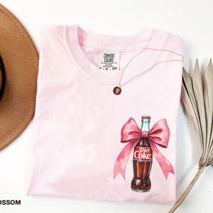 May include: Pink t-shirt with a graphic of a Diet Coke bottle with a pink bow. The shirt has a Comfort Colors tag.